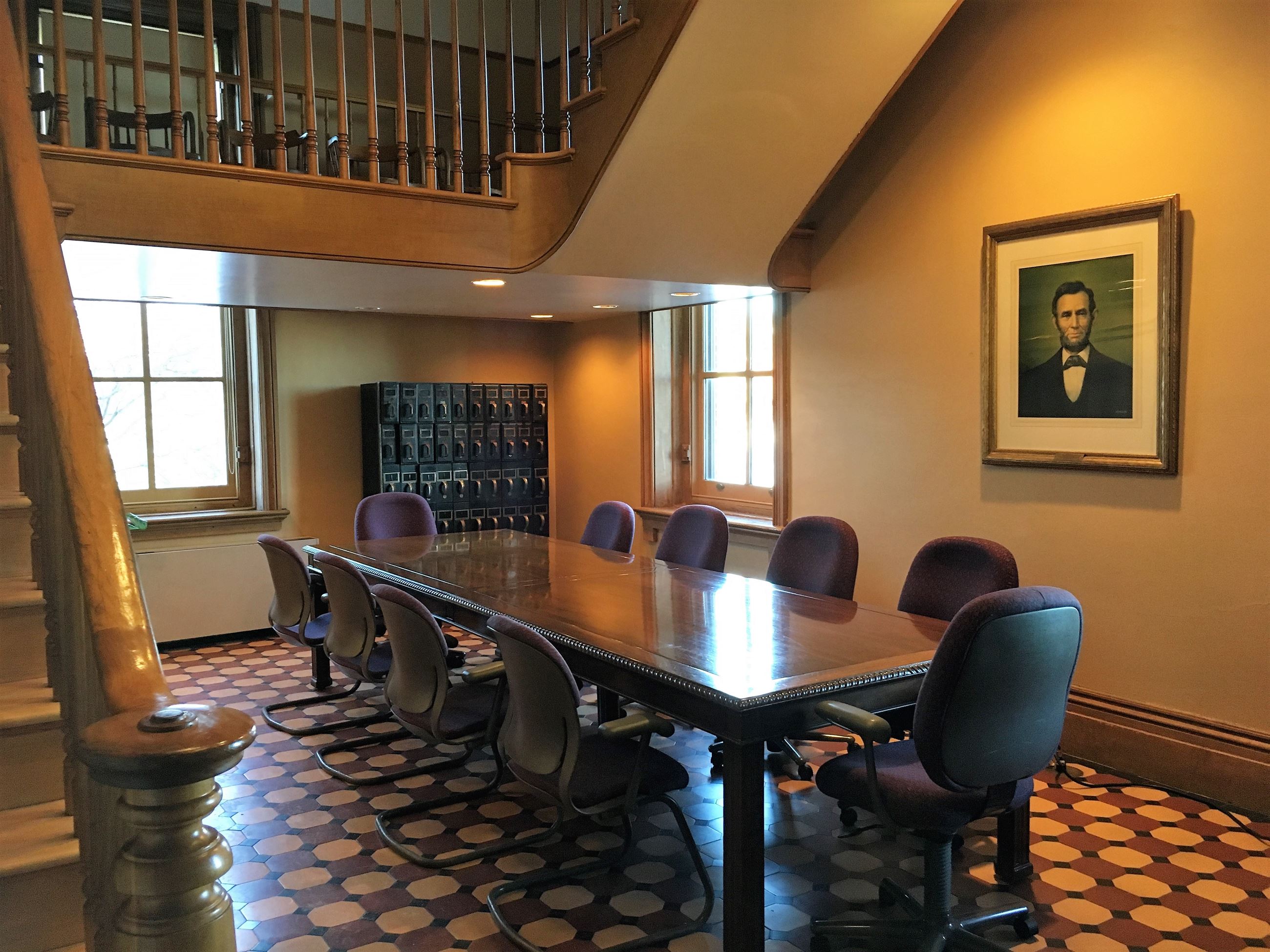 North Lobby Meeting Room with a staircase on the left and a large wooden table surrounded in the middle by office chairs. The walls are beige and the floor tiles are burgundy, tan, and black.