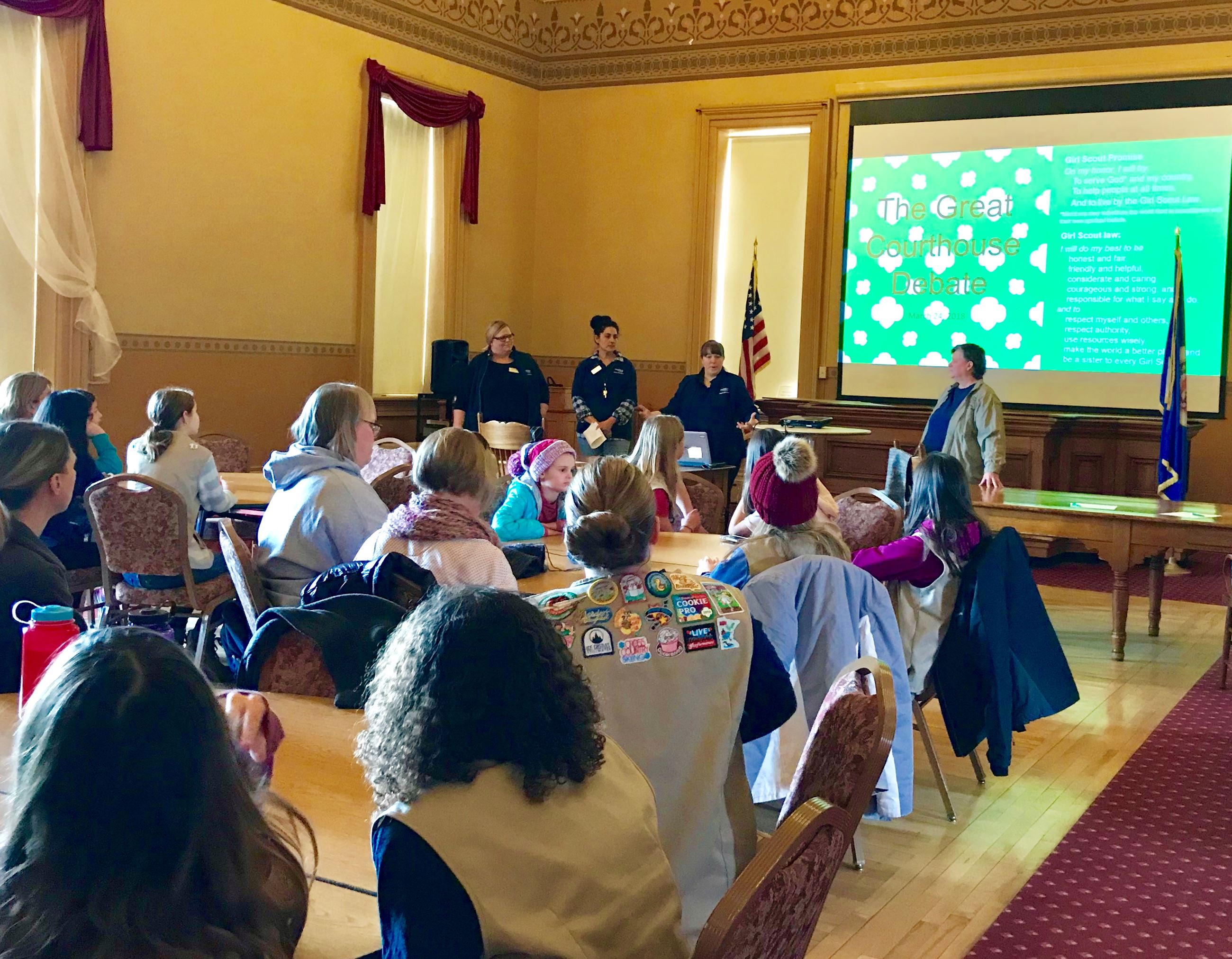 Girl scouts are seated at tables viewing a presentation on a large screen in the courtroom
