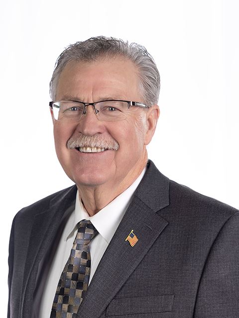 Portrait of a Commissioner Miron wearing glasses, a suit, tie, and a lapel pin, on white background.